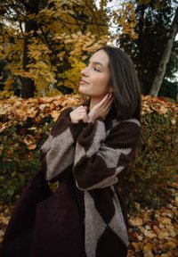 Femme portant un pull à motifs marron et beige, ajustant sa boucle d'oreille en plein air, avec des feuilles d'automne et des arbres en arrière-plan.
