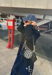 Donna che sistema un cappellino di denim blu, indossando un cappotto nero, occhiali da sole, pantaloni Adidas e una borsa a fantasia, in piedi su un pavimento piastrellato vicino a un allarme antincendio rosso.