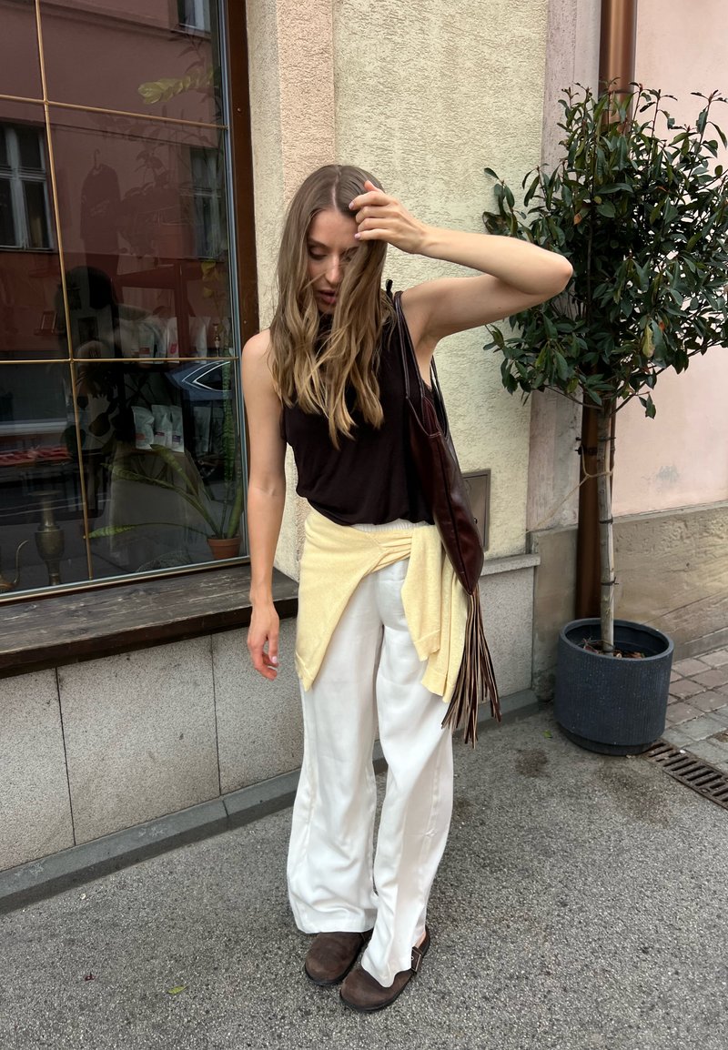 Jeune femme aux longs cheveux portant un haut noir, un pantalon blanc et des chaussures marron, se tient dehors près d'une fenêtre et d'une plante en pot, en ajustant ses cheveux.