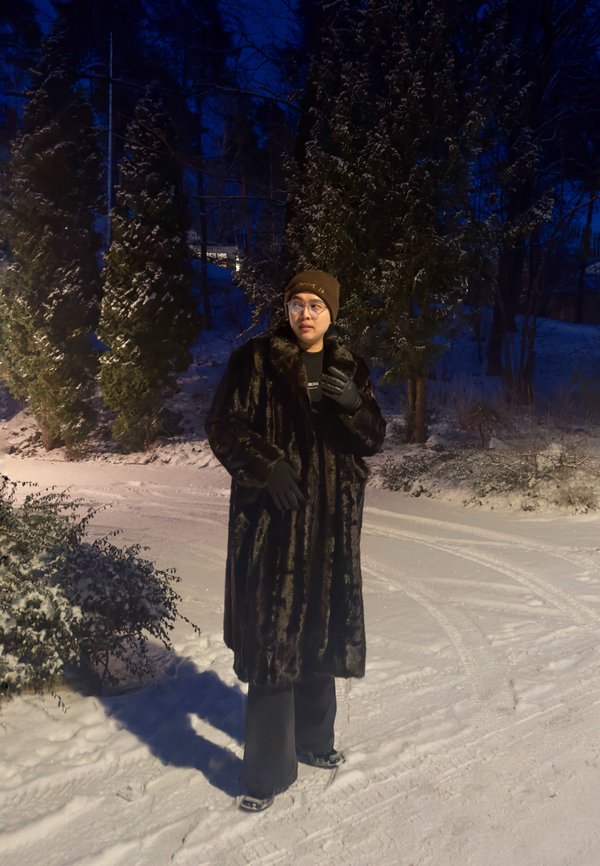 Personne portant un long manteau sombre, un chapeau marron, des lunettes et des gants, se tenant sur un sentier enneigé entouré de grands arbres la nuit.