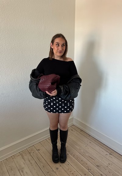 Joven mujer con top negro, falda de lunares, botas negras hasta la rodilla, sosteniendo un bolso tejido granate y chaqueta negra, de pie sobre un suelo de madera.