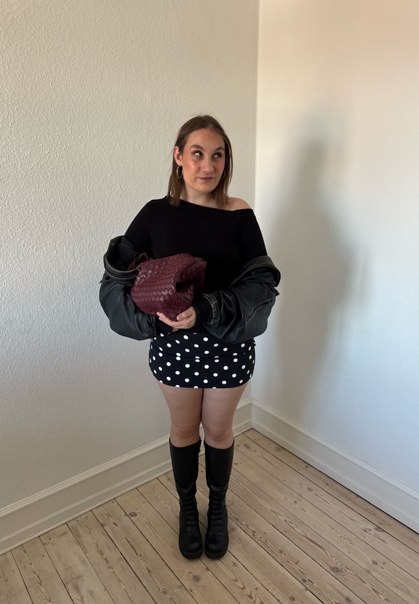 Jeune femme portant un haut noir, une jupe à pois, des bottes noires hautes jusqu'aux genoux, tenant un sac tissé bordeaux et une veste noire, debout sur un sol en bois.