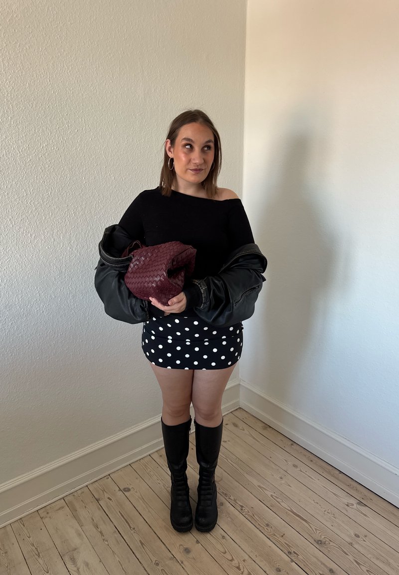 Junge Frau trägt schwarzes Top, gepunkteten Rock, kniehohe schwarze Stiefel, hält eine bordeauxrote gewebte Tasche und eine schwarze Jacke, steht auf Holzboden.