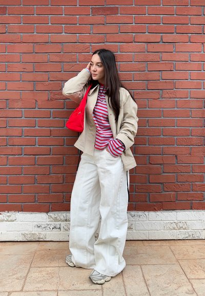 Mujer con chaqueta beige, camiseta de rayas rojas y blancas, pantalones anchos blancos, zapatillas grises y bolso rojo al hombro, de pie frente a una pared de ladrillos rojos.