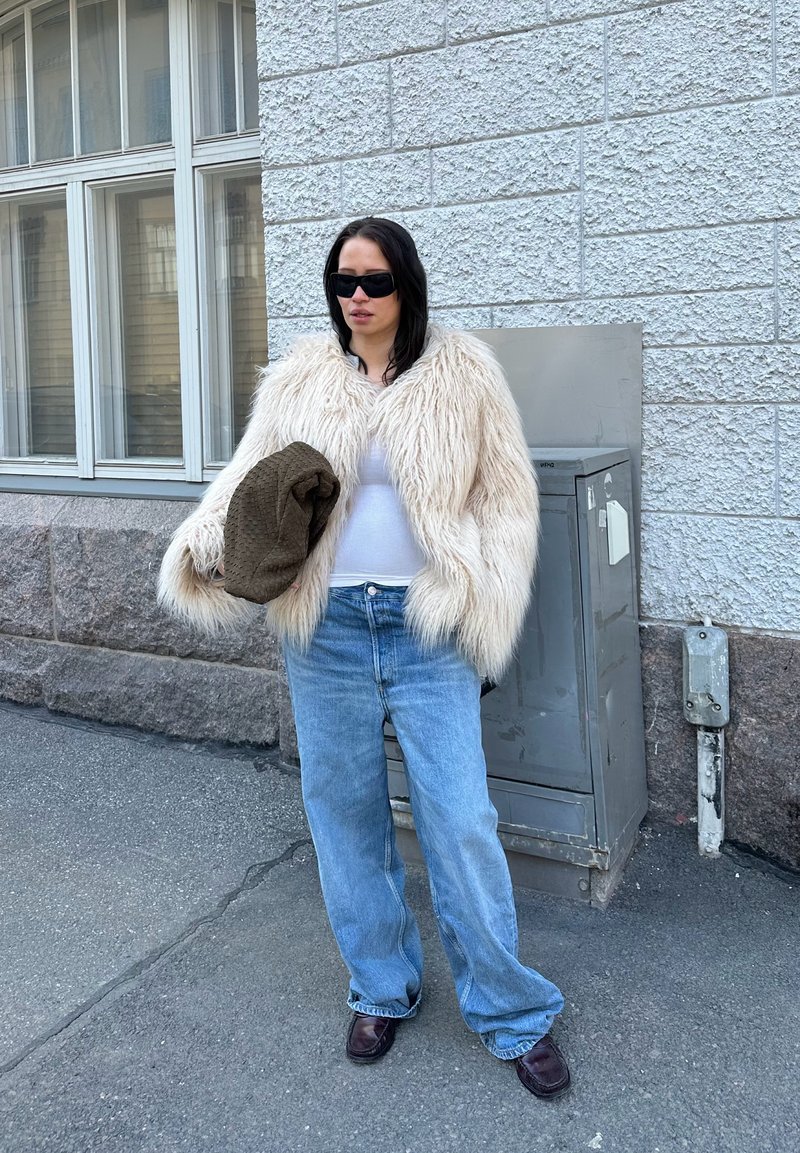 Femme en veste en fourrure beige surdimensionnée, top blanc, jean bleu ample et lunettes de soleil foncées se tient dans la rue près d'un bâtiment et d'une armoire utilitaire en tenant un sac marron.