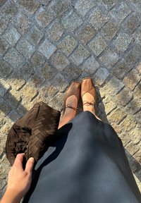 Sapatilhas de ballet em couro castanho com laços, uma saia escura e uma mala de tecido castanho escuro texturizado sobre uma superfície de calçada.