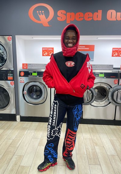 Sudadera roja con cuello alto, camisa negra debajo, y vaqueros con acentos azules que presentan gráficos en blanco y azul; calzando zapatos negros con acentos de color.