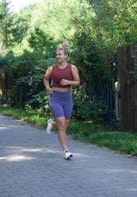 Atletiekoutfit: bordeauxrode crop top en paarse shorts. Lichte sneakers. Rennen op een verharde pad omringd door groen en bomen.