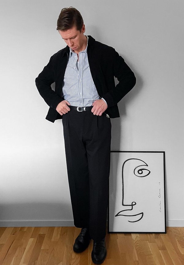 Veste noire, chemise à rayures blanches et bleues, pantalon noir et chaussures noires. Dessin géométrique remarquable encadré posé sur le sol.