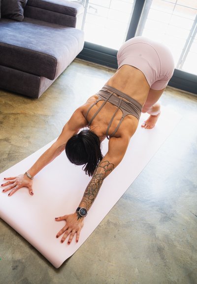 Alfombrilla de yoga en rosa pálido, persona con ropa deportiva beige realizando la postura del perro boca abajo sobre un suelo texturizado, tatuaje deportivo visible en el brazo.