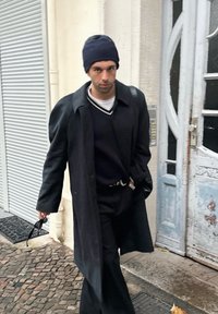 Čierny vlnený kabát, námornícka pletená čiapka, biela košeľa s čiernymi pruhmi, čierne nohavice. Drží slnečné okuliare, kráča pri svetlých dverách.