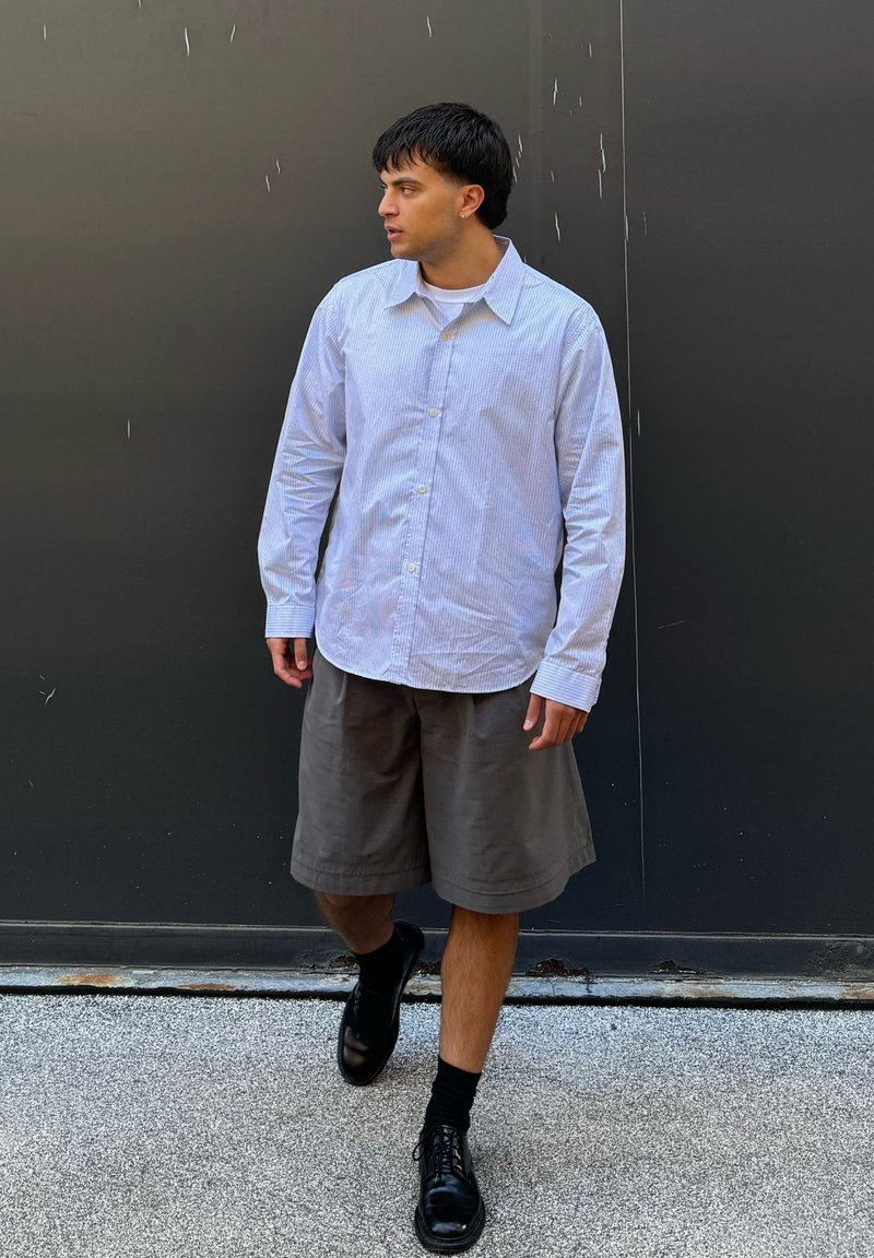 Chemise à manches longues bleu clair et blanche à rayures, associée à un short gris jusqu'aux genoux et des chaussures noires à lacets.