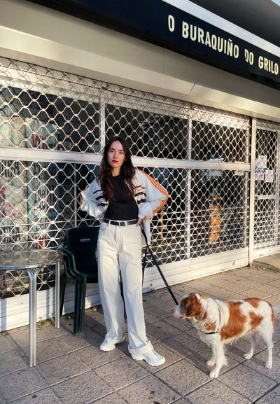 Chaqueta deportiva con rayas naranjas, parte superior negra y pantalones anchos blancos. Un perro con correa está de pie al lado de la persona sobre un pavimento texturizado.
