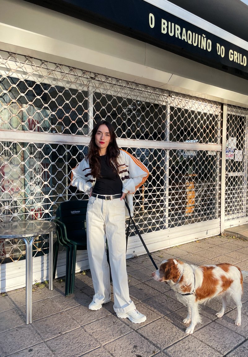 Αθλητικό μπουφάν με πορτοκαλί ρίγες, μαύρη μπλούζα και λευκά παντελόνια σε φαρδιά γραμμή. Ένα σκυλί με λουρί στέκεται δίπλα στο άτομο πάνω σε μια υφασμένη επιφάνεια.