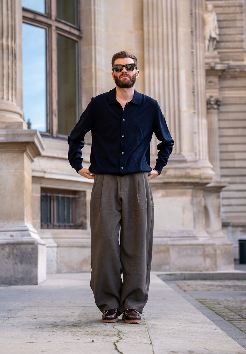 Homme barbu portant des lunettes de soleil noires et une chemise sombre à boutons avec un pantalon gris ample, debout devant un bâtiment en pierre de style classique.