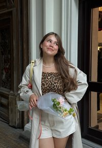 Mlada žena z dolgimi rjavimi lasmi, oblečena v bež trenčkot in leopardji vzorčast top, drži šopek mešanih rož in se smehlja.