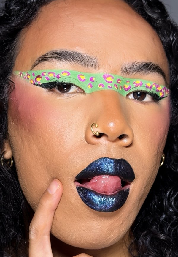 Personne aux cheveux bouclés portant un maquillage des yeux vert vif avec des taches roses, un rouge à lèvres métallique bleu foncé, un anneau au nez, et un doigt touchant son visage.