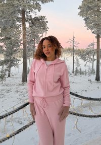Vrouw in een roze hoodie en broek met bruine handschoenen staat op een besneeuwd pad met verlichte touwlampen, omringd door met sneeuw bedekte bomen bij zonsondergang.