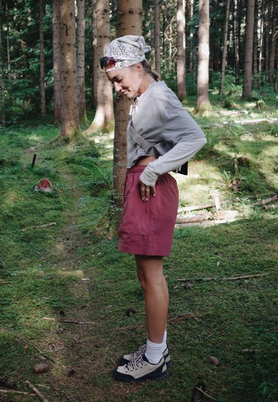Mujer con zapatos de senderismo, pantalones cortos y una camiseta de manga larga parada en un sendero del bosque rodeada de árboles y vegetación.