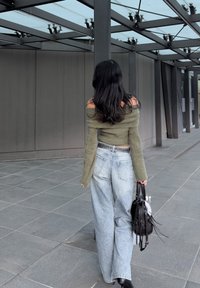 Pull olive green à épaules dénudées, jeans taille haute bleu clair avec une ceinture, sac à main noir et bottines noires sur une surface carrelée.