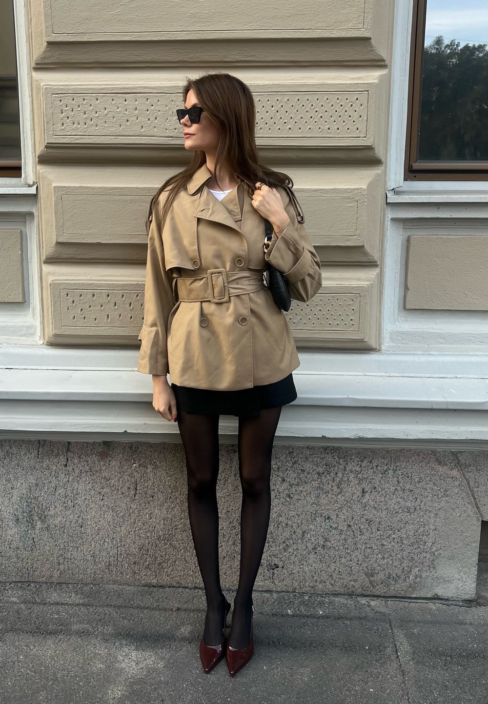 Beige trenchcoat med bälte i midjan, parat med en svart kjol och svarta strumpbyxor. Glansiga burgundy-höga klackar. Bakgrunden har en texturerad vägg.