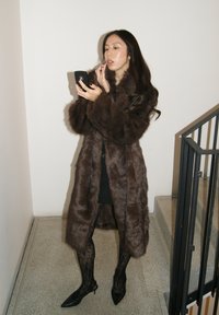 Abrigo marrón de piel sintética con cuello de chal, llevado sobre un vestido negro. Medias de encaje negras y tacones negros puntiagudos completan el conjunto. Enfoque en el teléfono.