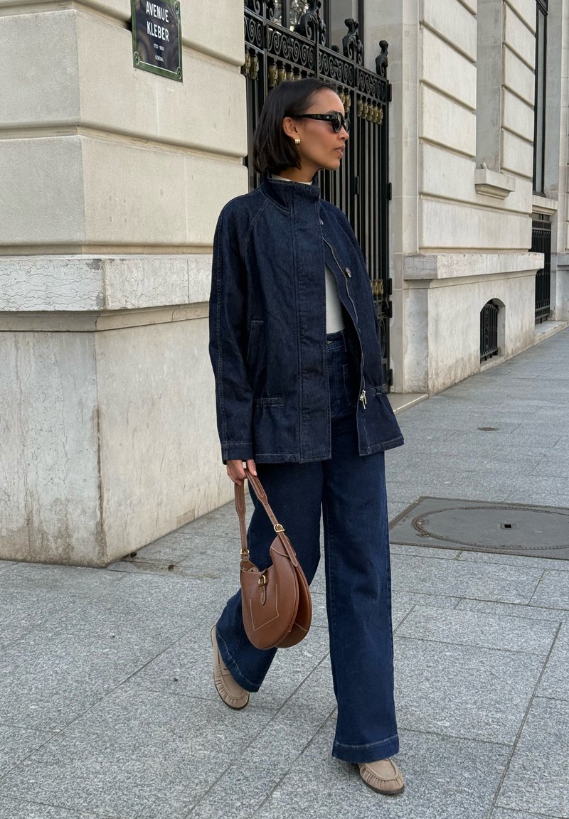 Femme portant une veste et un pantalon en denim foncé, chaussures beiges, portant un sac à main marron, marchant sur un trottoir en ville près d'un bâtiment en pierre et d'une grille métallique.