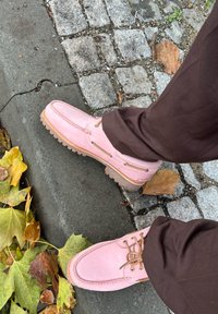 Sapatos de camurça rosa claro com atacadores castanhos e solas castanhas, apresentando um design de moccasin, posicionados numa calçada de pedras com folhas de outono.