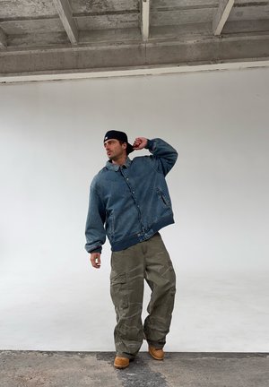 Hombre con chaqueta de mezclilla, pantalones cargo oliva, botas marrón claro y gorra negra puesta hacia atrás posa frente a una pared blanca bajo un techo de concreto.