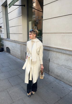 Abrigo beige con un cinturón, llevado sobre un suéter ligero y pantalones negros. Sostiene un bolso pequeño y lleva una bufanda ligera con flecos.