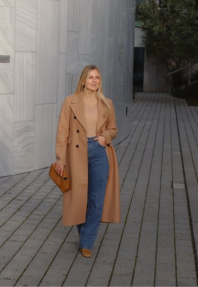 Abrigo de lana beige con botones de doble botonadura, suéter de cuello alto beige, jeans azules de tiro alto, clutch color tostado y mocasines marrones sobre un camino de piedra.