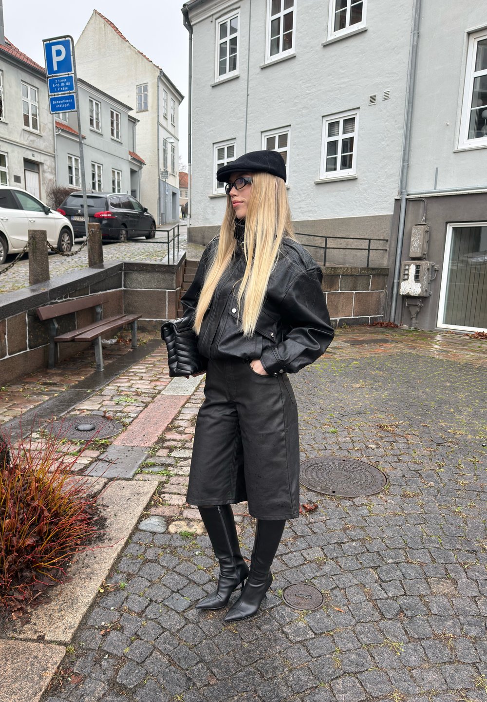 Veste aviateur en cuir noir portée sur un top noir ajusté et un short, assortie à des bottes noires pointues montant jusqu'aux genoux. Cheveux blonds longs, accessoires incluant une casquette noire.