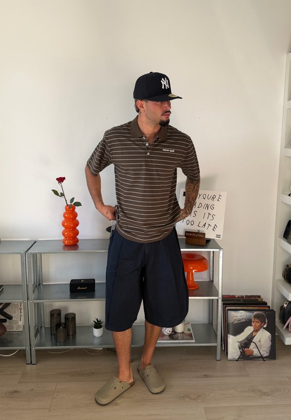 Homme portant un polo marron rayé, un short bleu marine, des chaussures beiges faciles à enfiler et une casquette noire, debout les mains sur les hanches dans une pièce moderne avec des étagères.
