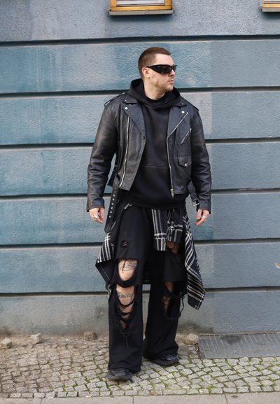 Chaqueta de cuero negra estilo motorista sobre una sudadera con capucha negra, combinada con pantalones negros sueltos con rotos y una superposición de cuadros. Gafas de sol completan el conjunto.