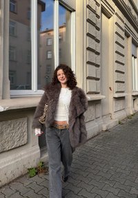 Manteau en fausse fourrure gris foncé avec de larges revers, porté sur un haut blanc et un pantalon gris clair. Porte un sac à main à motif en marchant.
