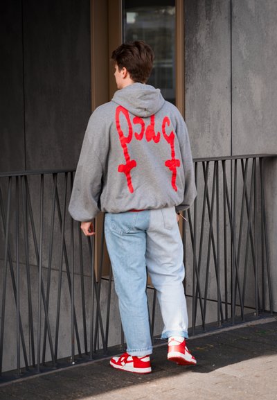 Sudadera gris con texto gráfico rojo y cruces en la parte posterior, combinada con jeans anchos de color azul claro y zapatillas rojas y blancas.