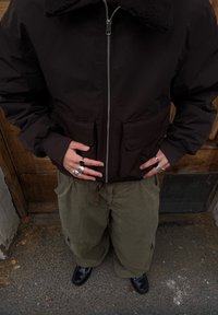 Persoon die een zwart jack met ritssluiting draagt, olijfgroene cargobroek, zwarte schoenen en meerdere zilveren ringen aan beide handen, die voor de taille zijn samengevouwen.