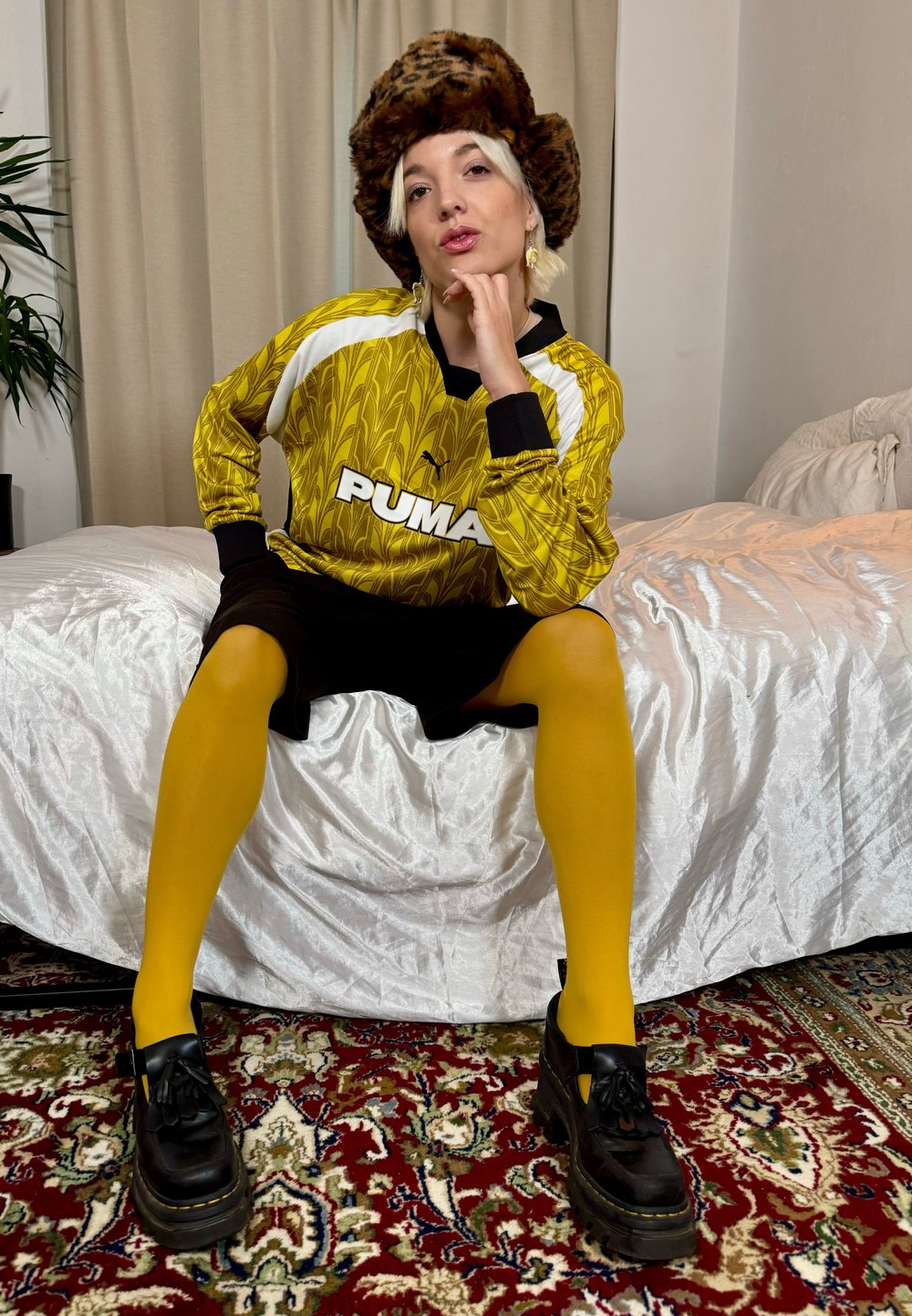 Geel gemarmerde sporttrui met zwarte accenten, zwarte shorts, felgele tights en zwarte platform schoenen, voor een witte dekbedovertrek.