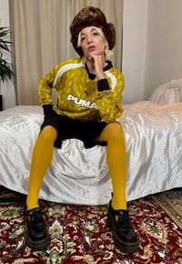 Geel gemarmerde sporttrui met zwarte accenten, zwarte shorts, felgele tights en zwarte platform schoenen, voor een witte dekbedovertrek.