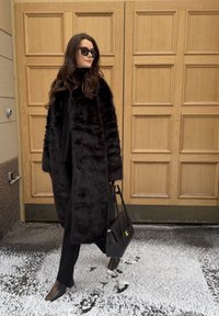 Femme portant un manteau en fourrure noir, un pantalon noir, des bottes et des lunettes de soleil, tenant un sac à main noir, debout sur un sol enneigé près d'une porte en bois.