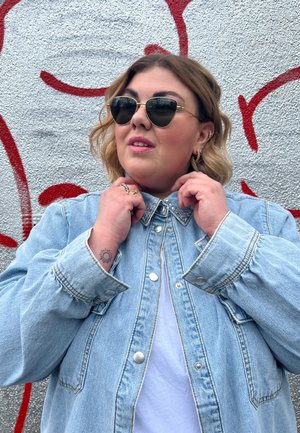 Chaqueta de denim azul claro con un diseño estructurado, cuello adornado y botones a presión plateados. El modelo lleva gafas de sol oscuras y una camiseta blanca.