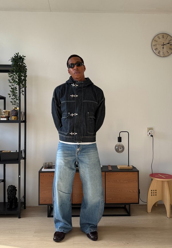 Un homme portant des lunettes de soleil, une veste noire à brandebourgs, un jean bleu ample et des chaussures sombres se tient dans une pièce meublée.