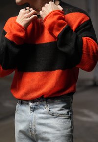 Μαύρος και πορτοκαλί ριγέ πλεκτός πουλόβερ με μακριά μανίκια, φορεμένο πάνω από ανοιχτό μπλε τζιν. Το πουλόβερ έχει χαλαρή εφαρμογή και υφή υφάσματος.