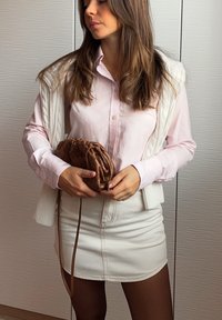 Lichtroze button-up shirt onder een crèmekleurige gebreide vest, gecombineerd met een witte denimrok. Bruine gehaakte handtas voor zich gehouden.