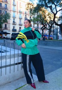 Femme en veste verte et noire avec des rayures jaunes, pantalon noir avec des rayures blanches, et chaussures rouges pointues posant en plein air dans un cadre urbain.