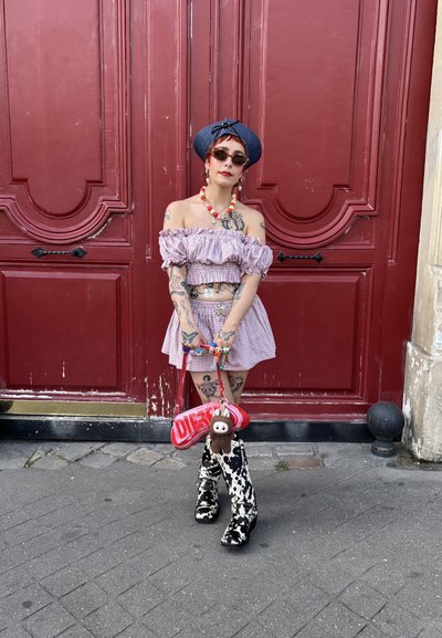 Top lila sin mangas, con volantes, falda a juego, botas de estampado de vaca, bolso rojo con llavero de peluche, collar de cuentas coloridas y gafas de sol.