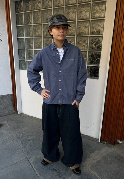 Joven con camisa azul marino abotonada, pantalones negros de pierna ancha, gorra de camuflaje y anillos, de pie con una mano en la cadera junto a una pared de bloques de vidrio.