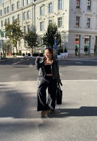 Zwarte leren jack over een zwarte crop top, wijde zwarte broek en bruine laarzen, met een zwarte tas in de hand, staande op een zonnige straat.
