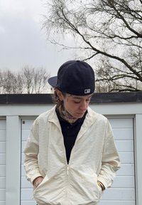 Light cream zip-up jacket with a subtle embossed pattern, worn over a black polo shirt, complemented by a navy baseball cap.