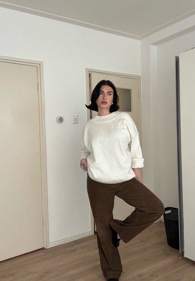 Mujer con cabello negro, vestida con un suéter blanco y pantalones marrones, de pie en interiores con una pierna levantada, manos en los bolsillos, frente a paredes blancas y puertas cerradas.